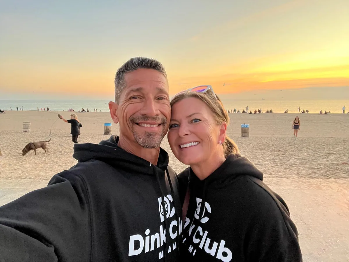 Heather and Marcelo Reyes, owners of Dink Club, on the beach in Miami at sunset wearing Dink Club hoodies